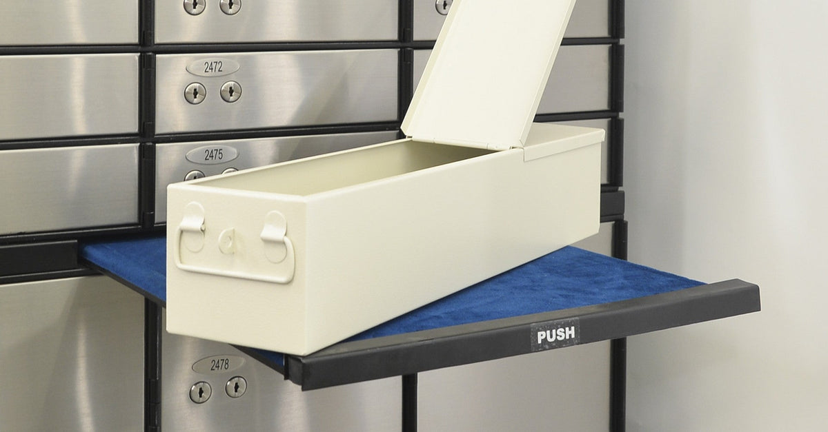 Open cream-colored safety deposit box drawer with raised lid on a blue counter in front of a bank safety deposit locker wall.